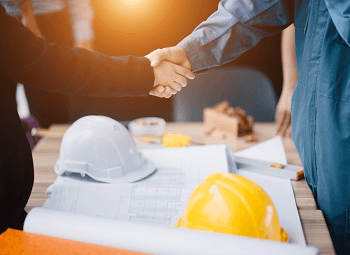 Two people shaking hands over a table that has blueprints and hard hats on it