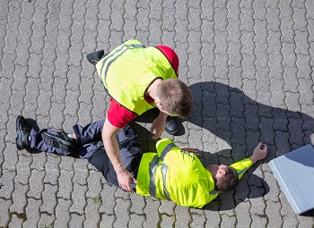 Two workers wearing reflective vests, one is checking on the other who has collapsed on the ground